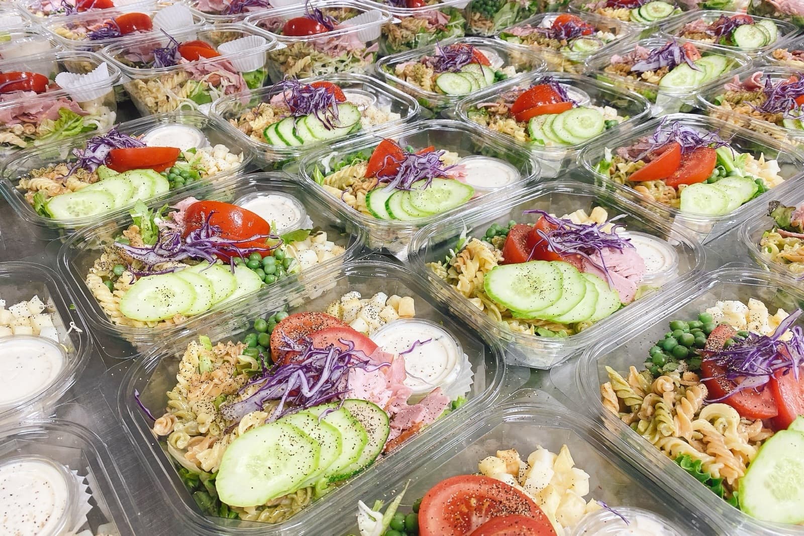 포장을 마친 신선식이 작업대 위에 줄지어 놓여 있다. 각 용기에 오이, 방울토마토, 적양배추, 파스타, 완두콩과 드레싱 컵이 담겨 있다