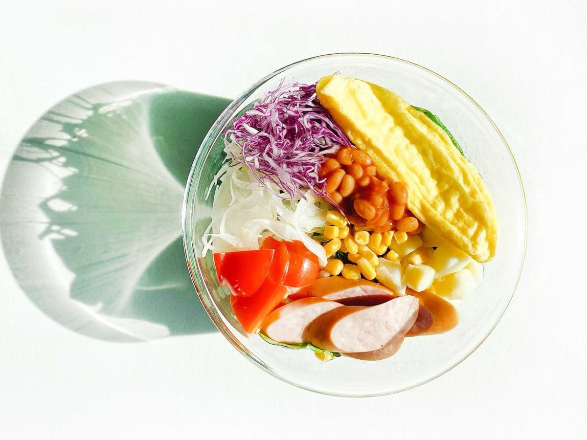 투명 볼에 담긴 샐러드밀. 오믈렛, 소시지, 베이크드빈, 옥수수, 방울토마토, 적양배추, 양파, 잎채소가 담겨 있다