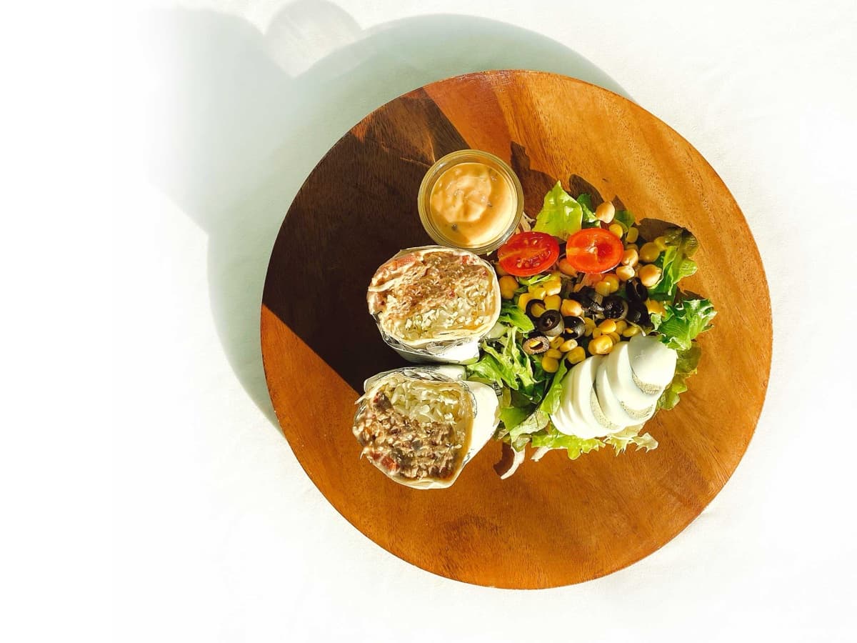 나무 도마에 놓인 반으로 자른 랩과 콥샐러드. 옥수수, 병아리콩, 블랙올리브, 방울토마토, 메추리알, 잎채소와 드레싱 컵이 함께 있다